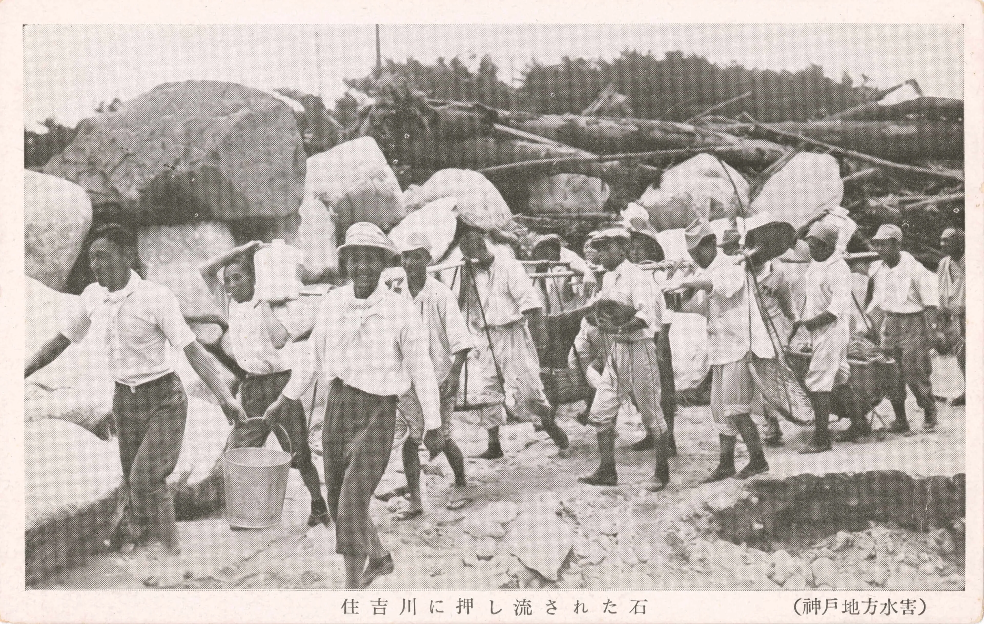 1938年に起きた阪神大水害で、大きな岩が押し流されてきて石垣のように積みあがっている。その脇を復興作業にいそしむ人々が歩いている様子を収めた写真絵はがき。
